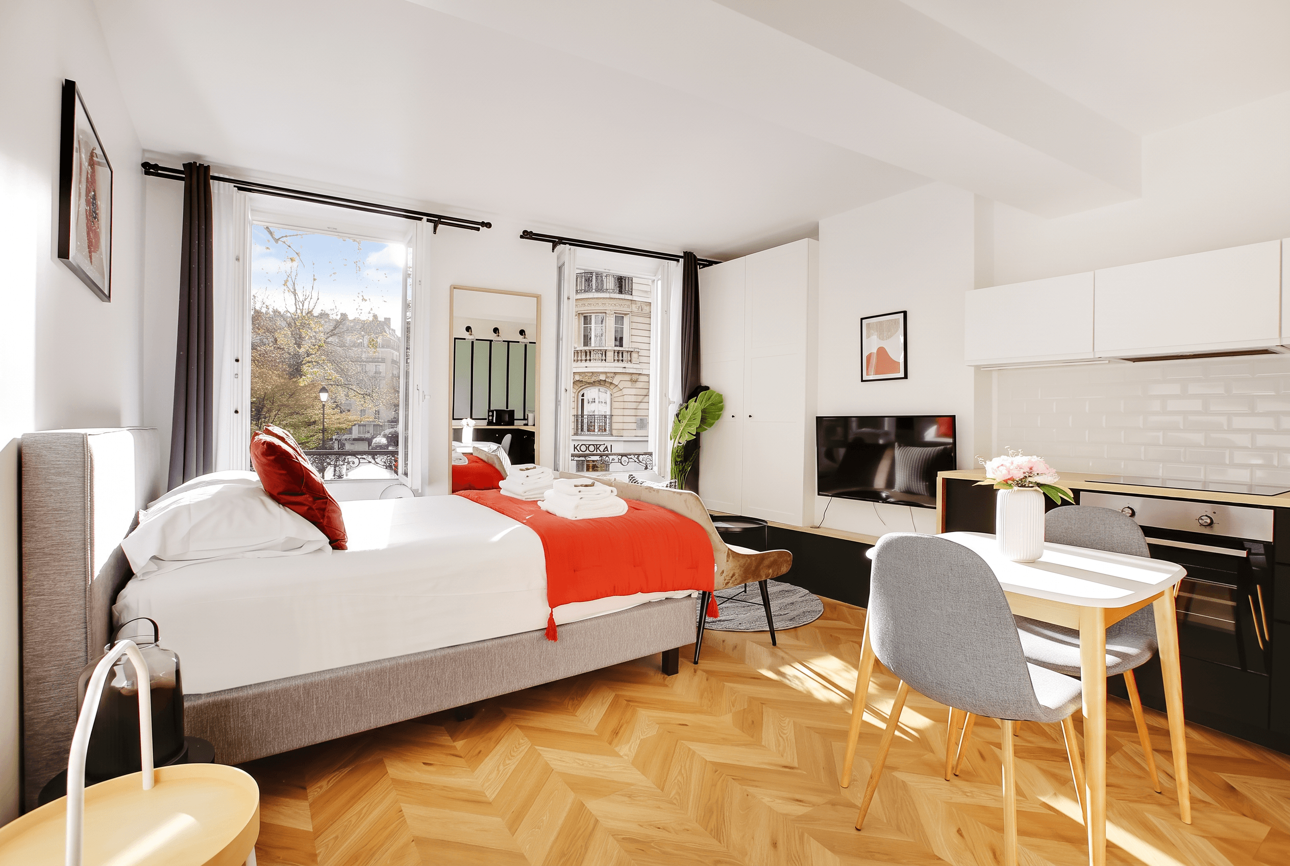 Intérieur d’un studio lumineux : parquet en chevrons, lit au linge rouge, table et chaises grises, grandes fenêtres