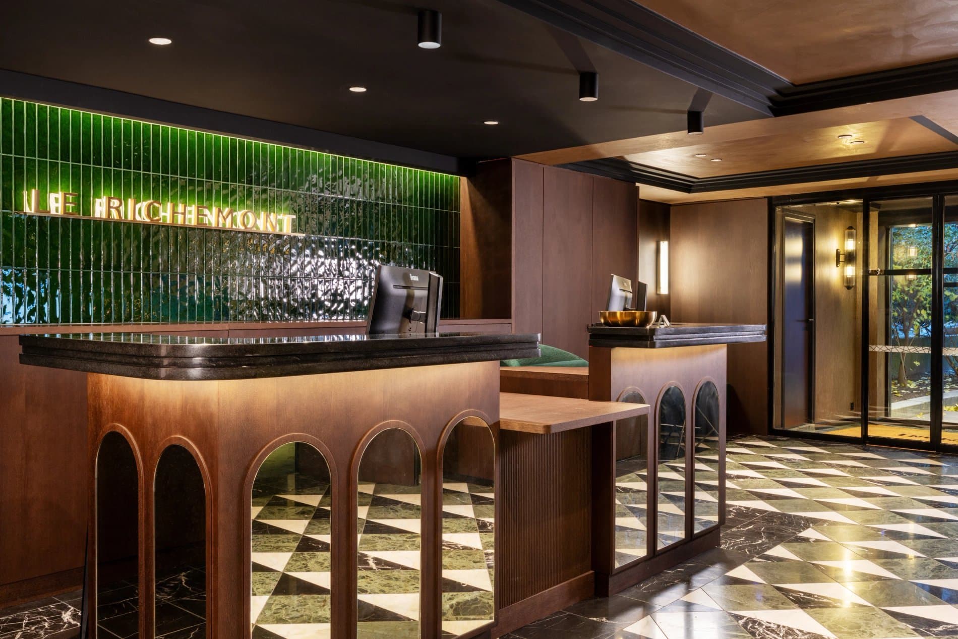 Hall d’accueil de l’Hôtel Le Richemont, avec son comptoir en bois précieux, son mur de carreaux vert émeraude et son enseigne lumineuse